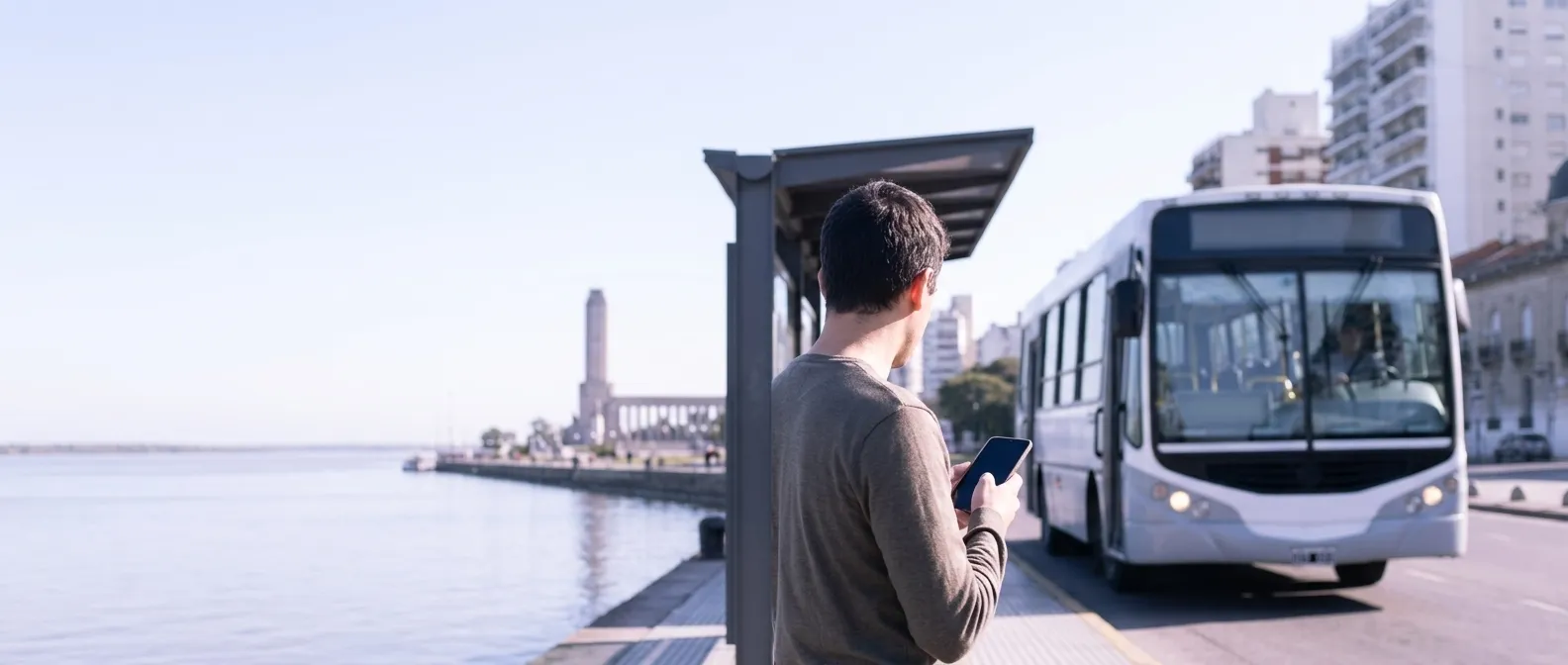 Una persona espera en una parada frente al río, con el teléfono en la mano, mientras llega un colectivo.
